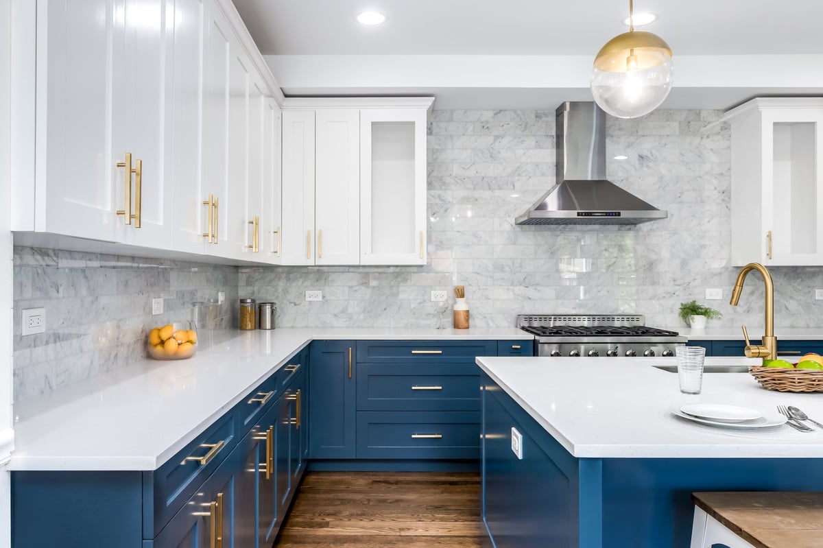 A luxurious white and blue kitchen with gold hardware, stainless steel appliances, marble subway tile backsplash, and white marble granite countertops