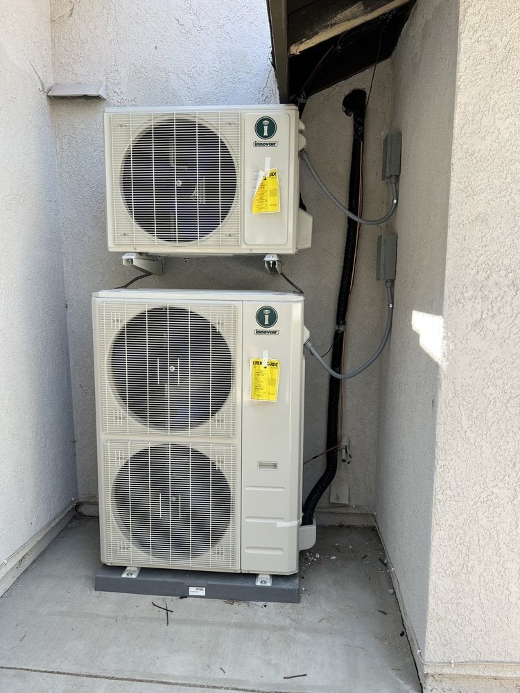 Two stacked air conditioning outdoor units with circular fan vents and yellow warning labels mounted on an exterior wall in a narrow alcove