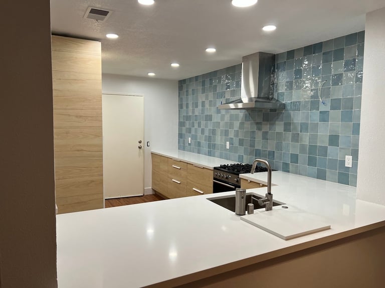 Modern kitchen with white countertop, blue tile backsplash, stainless steel range hood, and wood cabinetry.