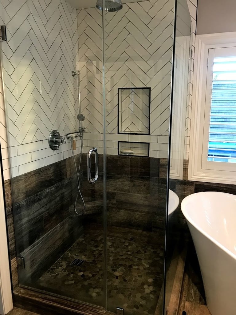 Modern bathroom with glass-enclosed walk-in shower featuring herringbone tile walls, dark wood-look tile base, and river rock floor