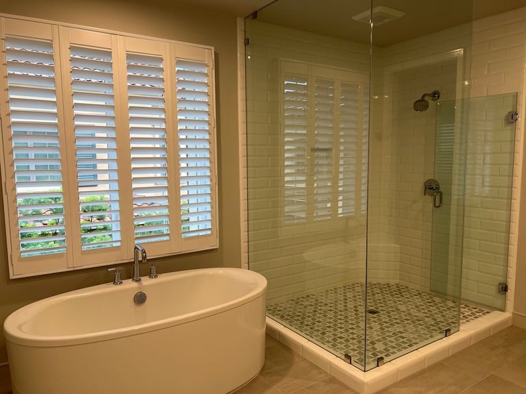 Modern bathroom with freestanding oval bathtub on left, walk-in glass shower on right, and shuttered windows overlooking exterior