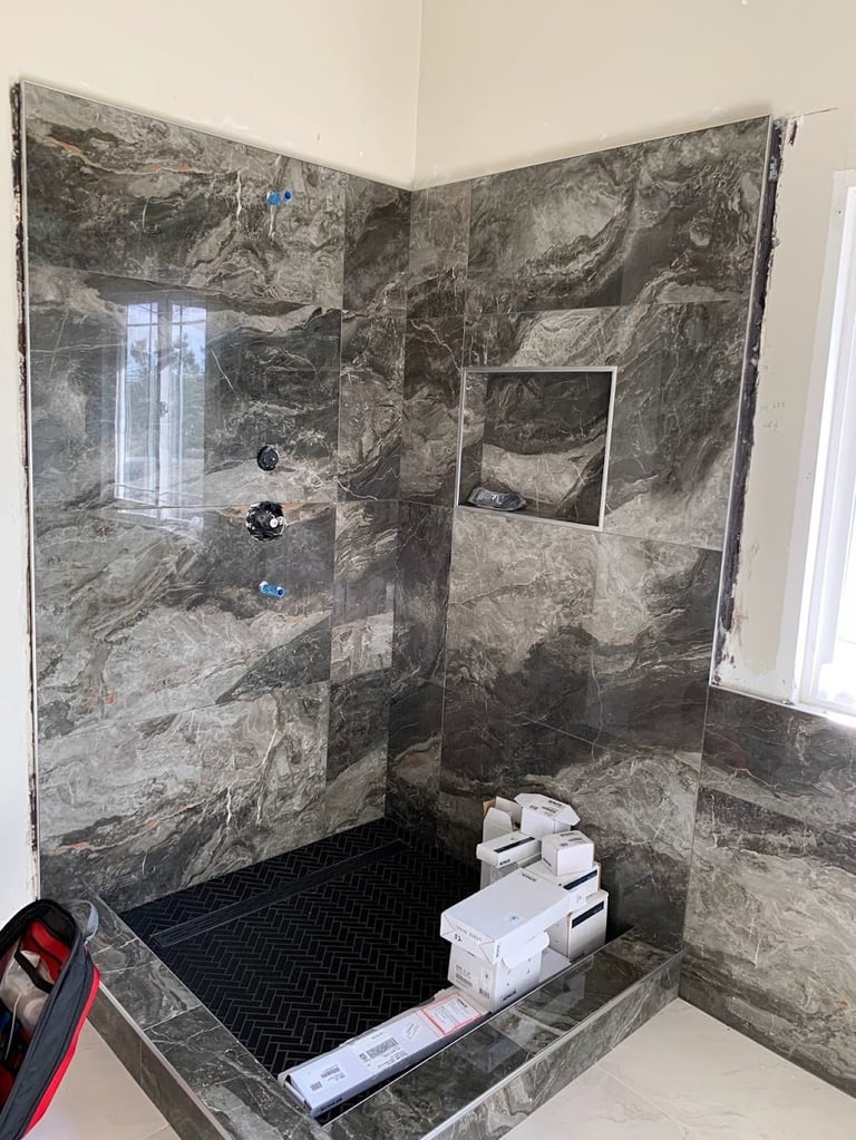 Modern bathroom shower with dark gray marble tile walls, glass enclosure, black mosaic floor, and white towels on built-in shelf