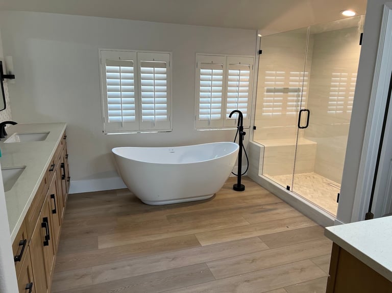 Modern bathroom with freestanding white bathtub, glass shower enclosure, wooden vanities, and white shuttered windows