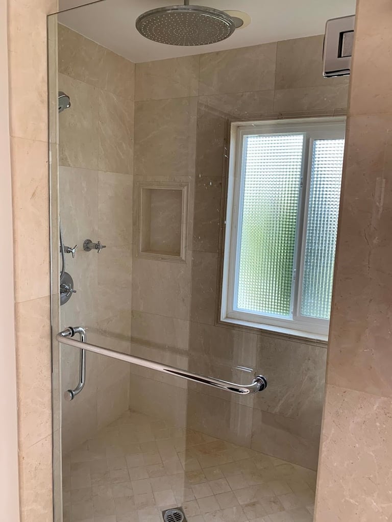 Modern walk-in shower with glass enclosure, tile walls, rainfall showerhead, and window overlooking greenery